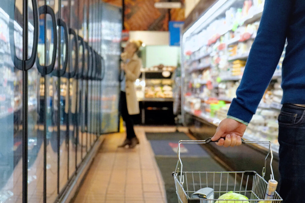 A shopper holding a grocery basket in a supermarket aisle, emphasizing the challenge of decoding food labels and avoiding misleading marketing claims.